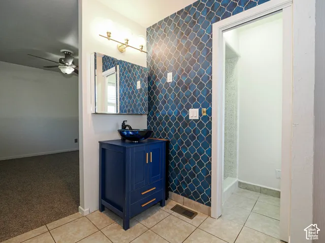 Blue vanity cabinet with gold-tone hardware, dark glass vessel sink, three-light wall fixture, and decorative patterned wallpaper accent wall