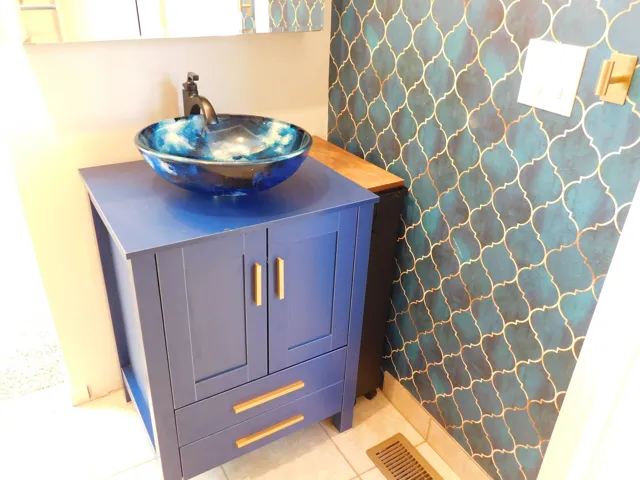 Vessel sink with a matte black faucet, blue vanity with gold-tone hardware, and decorative wall covering