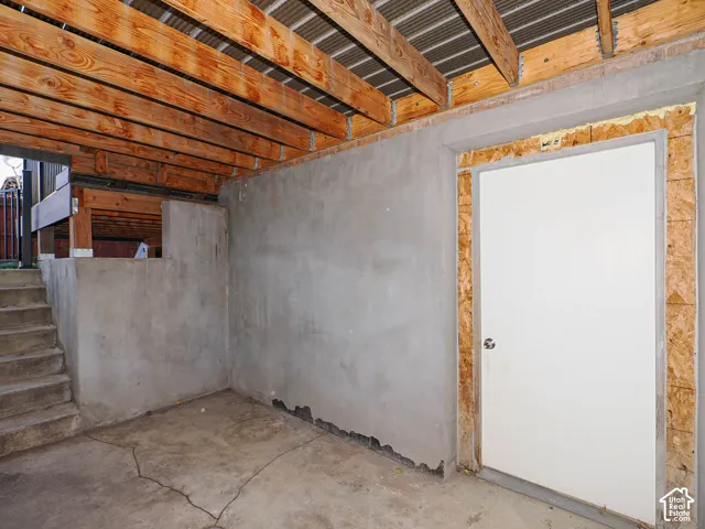Unfinished space featuring exposed timber joists, concrete flooring, cement-plastered walls, and a single-panel door with unfinished trim
