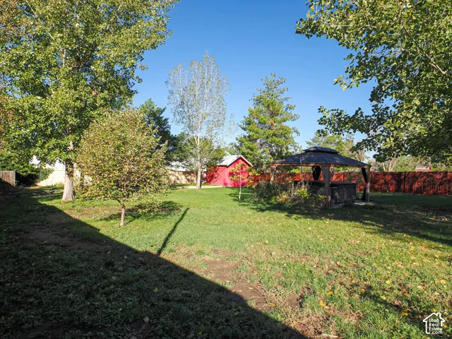 Expansive backyard featuring a manicured lawn, mature trees, a permanent metal-framed gazebo, and a detached storage structure