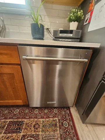Stainless steel dishwasher, light wood-finish cabinetry, white countertop, and subway tile backsplash