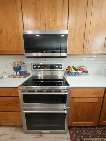 Stainless steel microwave and double oven, wood-finish cabinetry, light-toned countertop, and white subway tile backsplash
