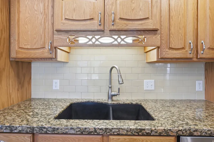 Light wood-finish cabinetry with decorative hardware, undermount double basin sink with pull-down faucet, granite-style countertops, and subway tile backsplash