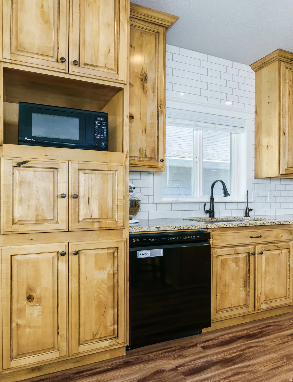 Wood-finish cabinetry, granite countertops, subway tile backsplash, black dishwasher, and wood-finish flooring