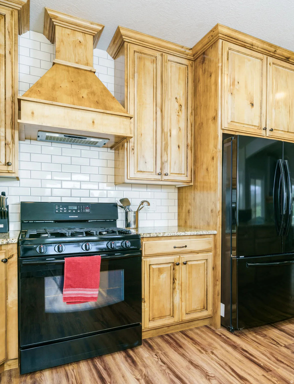 Kitchen featuring wood-finish flooring, light wood cabinetry, white subway tile backsplash, and granite-style countertops