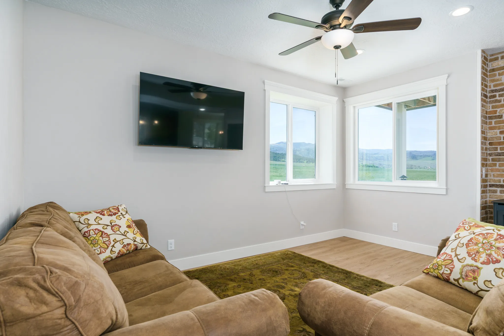Recessed ceiling lighting, wood-finish flooring, two large windows with white trim, ceiling fan with integrated lighting, and a brick-finish wall