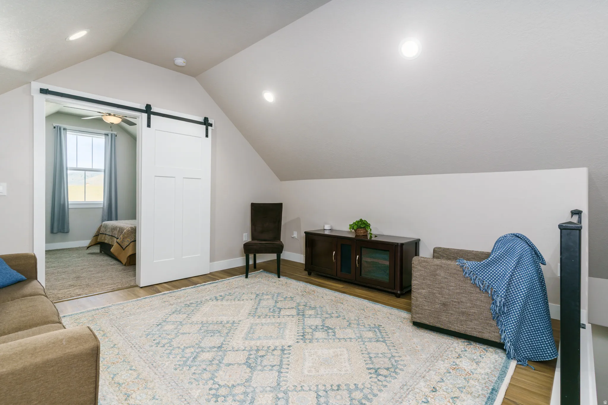 Sloped ceilings with recessed lighting, wood-finish flooring, white barn door with black hardware, and a window with a ceiling fan in the adjoining room