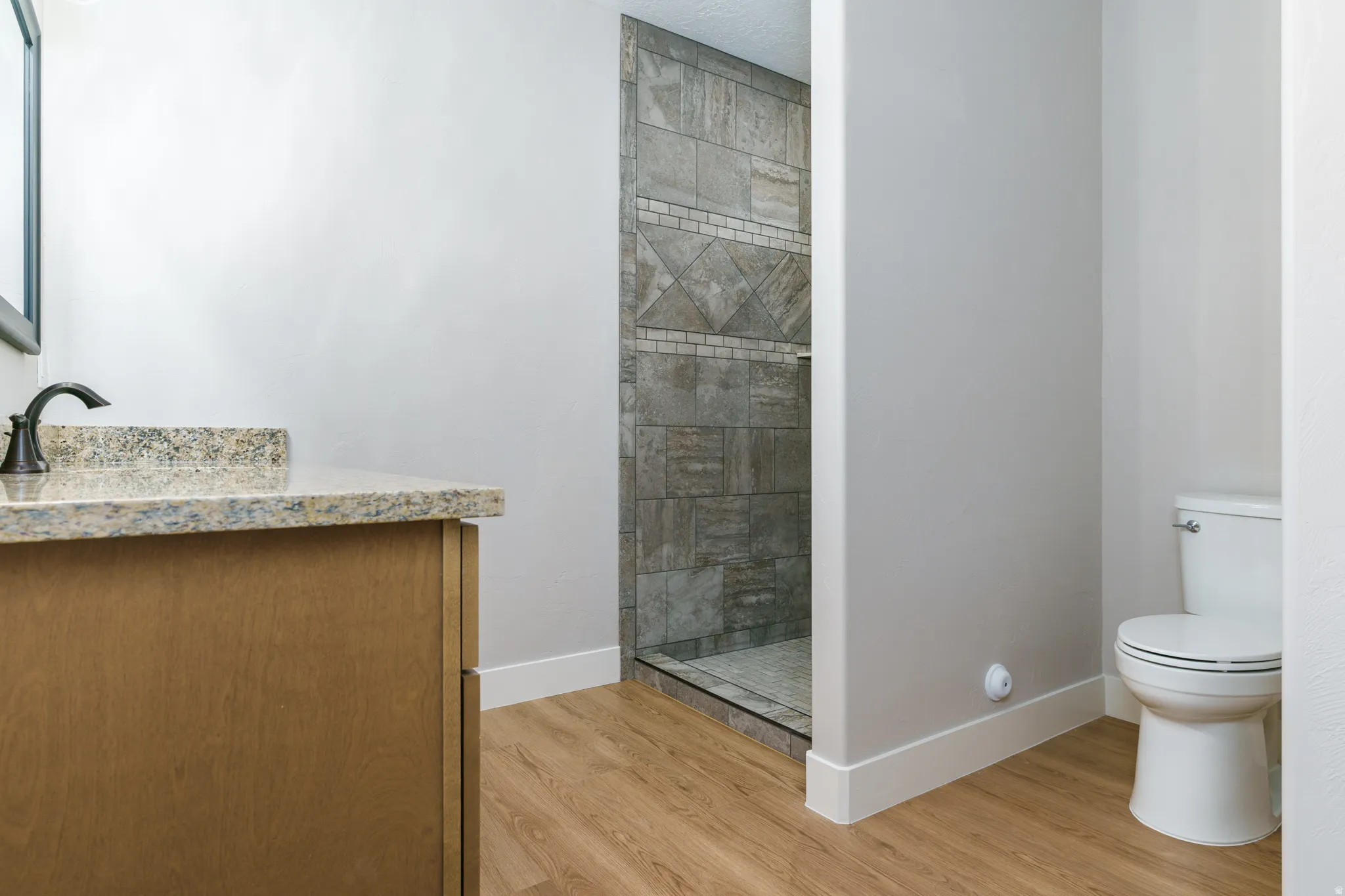 Wood-finish flooring, vanity with stone countertop, dark bronze faucet, and a tiled shower with glass enclosure