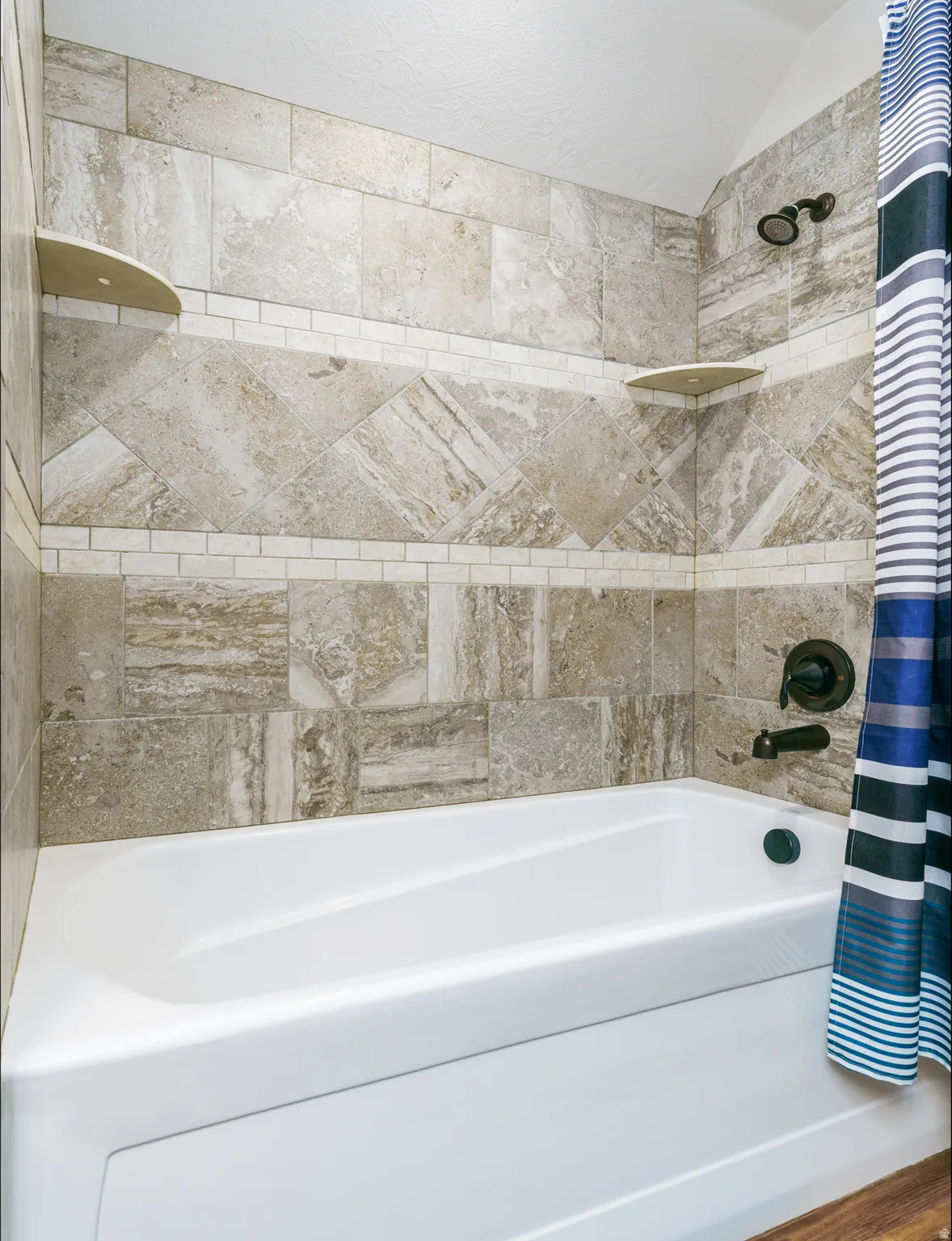 White bathtub with a bronze showerhead and fixture, set against a tiled wall featuring a decorative accent band and two corner shelves