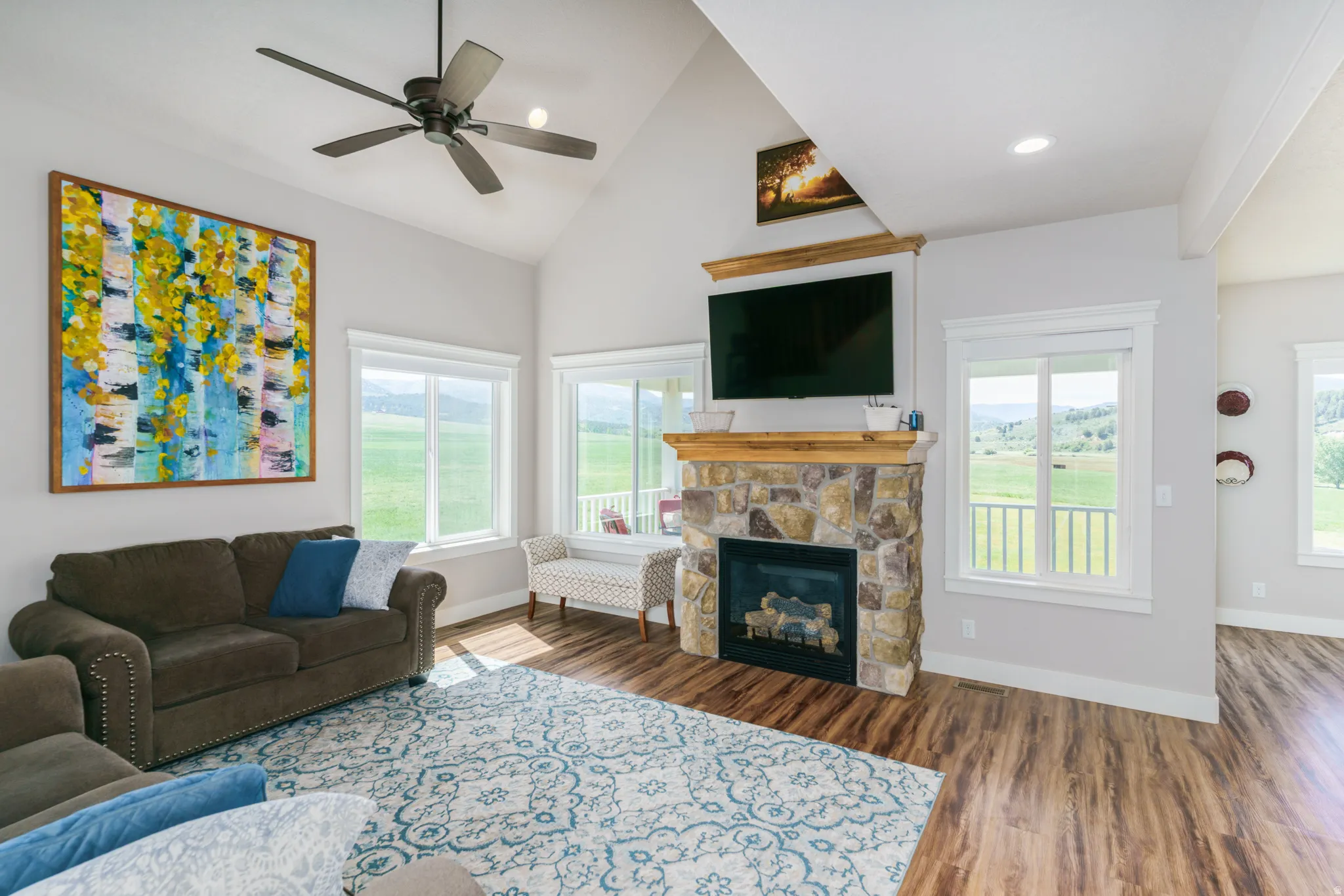 Vaulted ceiling with a ceiling fan, stone-surround fireplace with a wood mantel, wood-finish flooring, and multiple windows