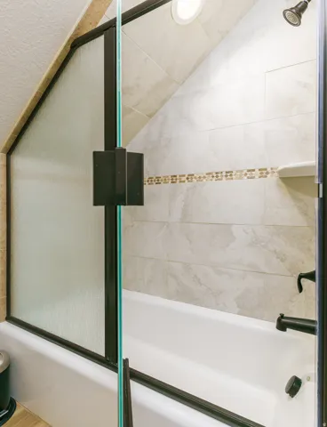 Bathtub with a glass shower enclosure, bronze-finish shower fixtures, and light-toned tile surround with a mosaic accent strip