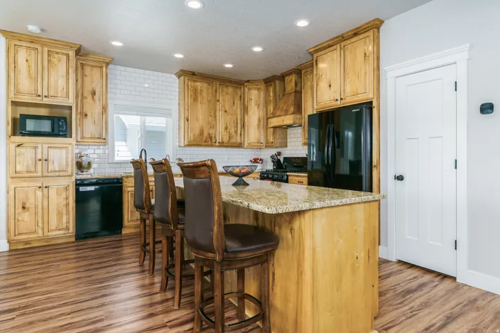 Wood-finish flooring, recessed lighting, subway tile backsplash, light wood cabinetry, and a kitchen island with a stone countertop