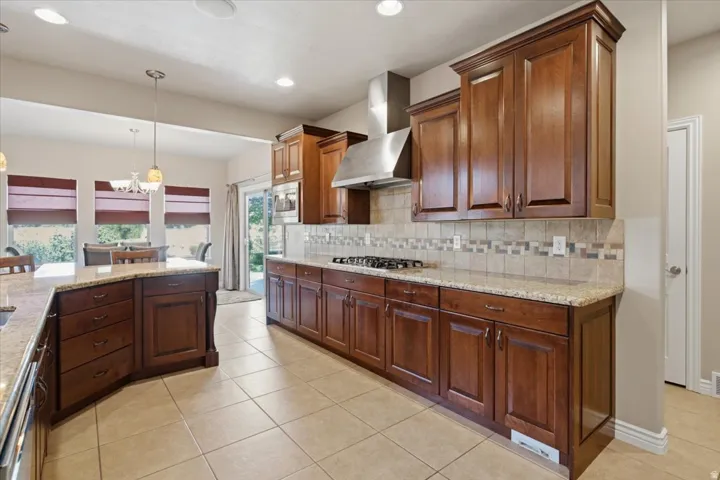 Kitchen featuring wood-finish cabinetry, granite-style countertops, stainless steel range hood, tile flooring, and a tile backsplash