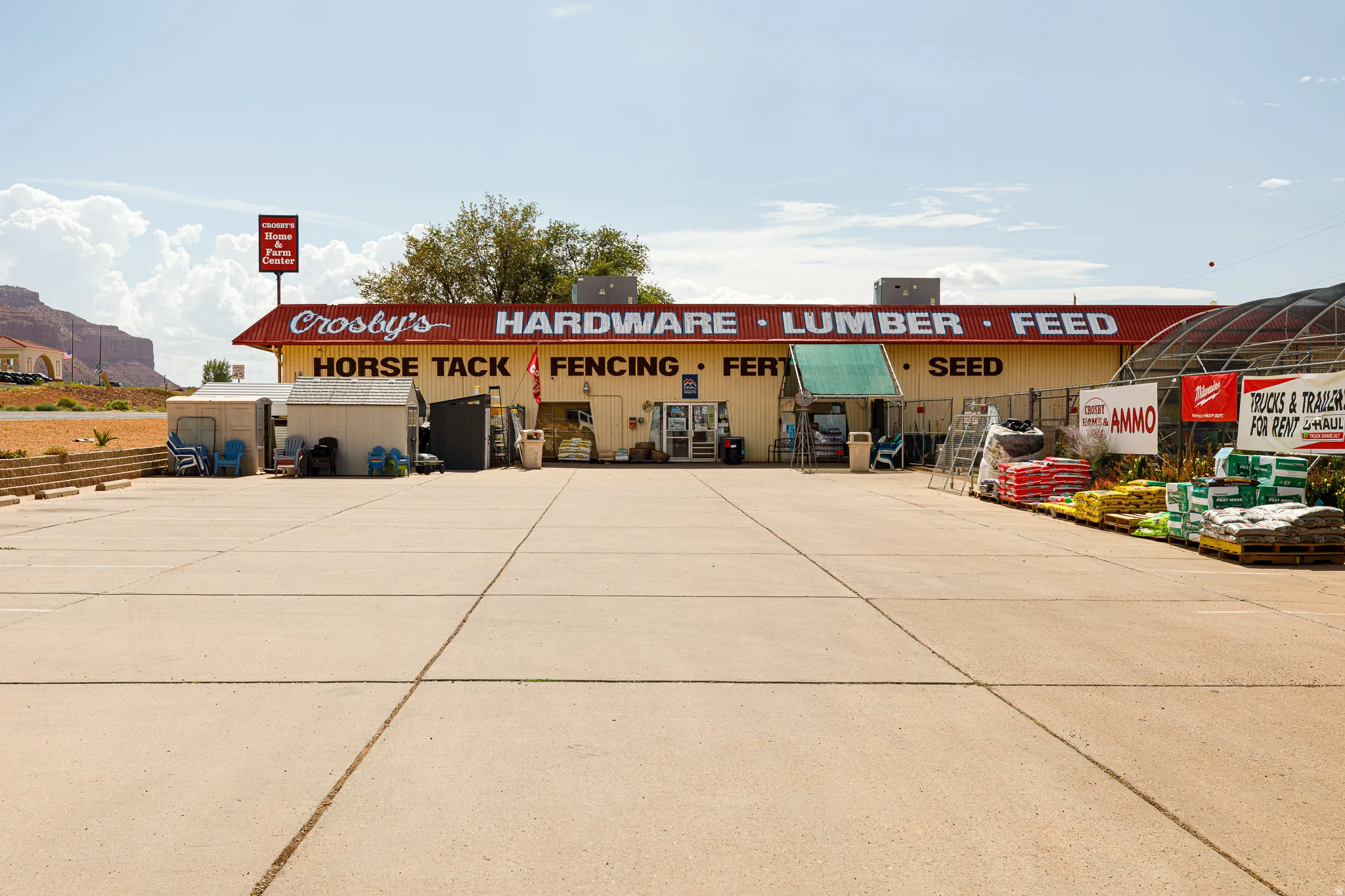 Expansive paved parking lot fronting a commercial structure with a red roof, a separate signage structure, and a covered outdoor retail area