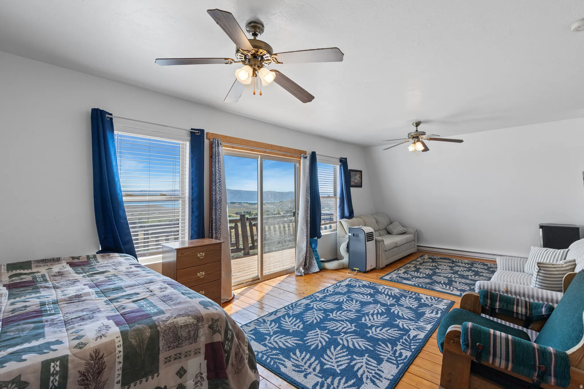 Bright, sun-filled bedroom with dual ceiling fans, wood-finish flooring, and sliding glass doors opening to an expansive deck with panoramic outlooks
