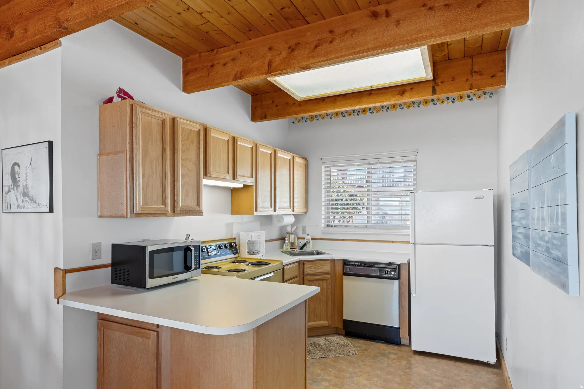 Bright kitchen featuring wood cabinetry, a centered island with prep space, stainless steel appliances including a dishwasher and refrigerator, skylight for natural illumination, and exposed wood beam ceiling detail