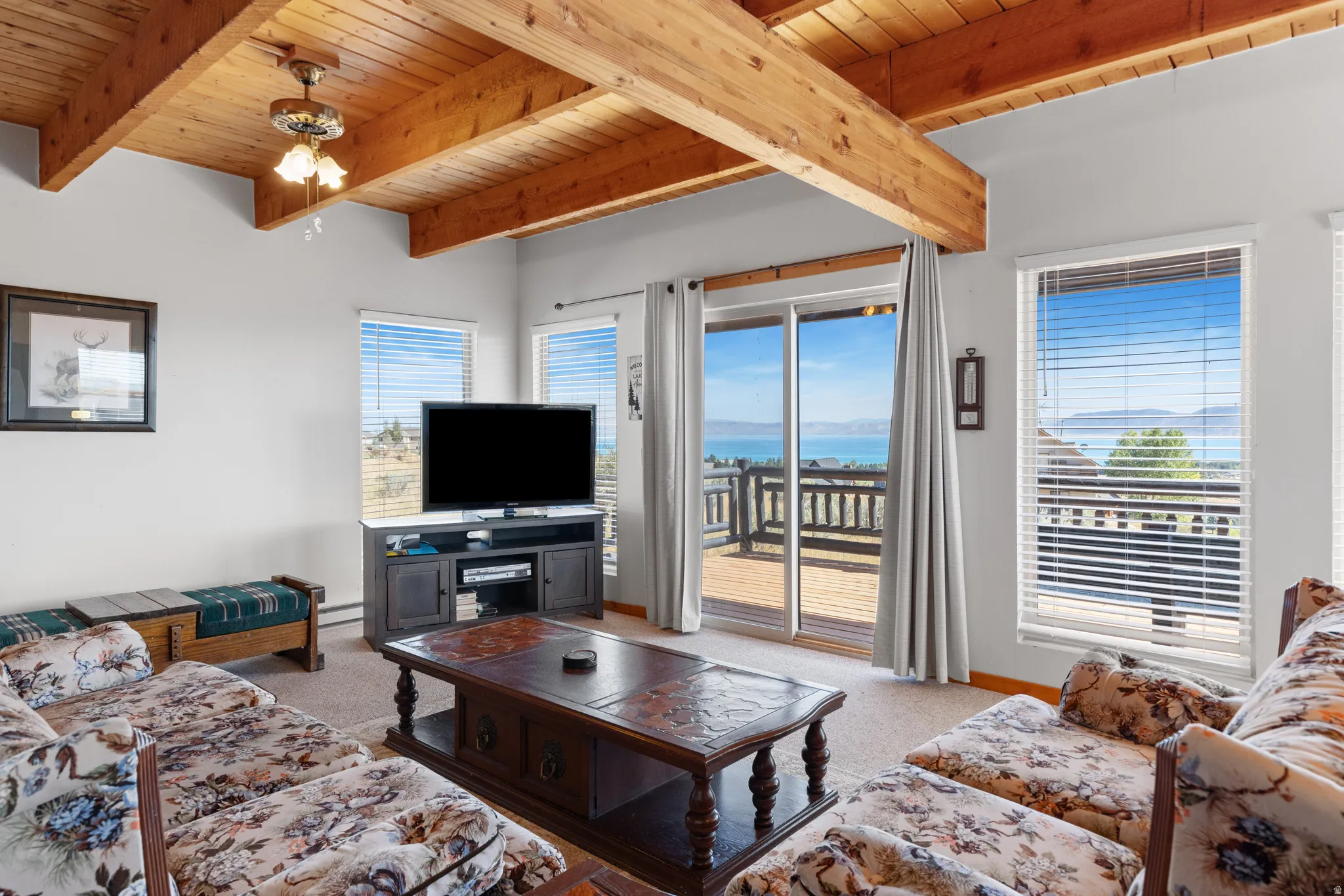 Exposed wood beam ceiling, ceiling fan with light fixture, sliding glass door to exterior deck, multiple windows with blinds, and carpeted flooring