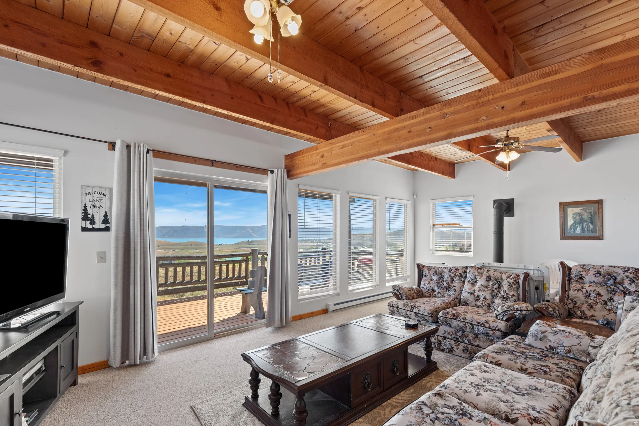 Cathedral wood-beam ceiling with exposed wood planks, large sliding glass door with exterior deck, multiple windows, built-in stove pipe, and recessed ceiling fan light fixtures