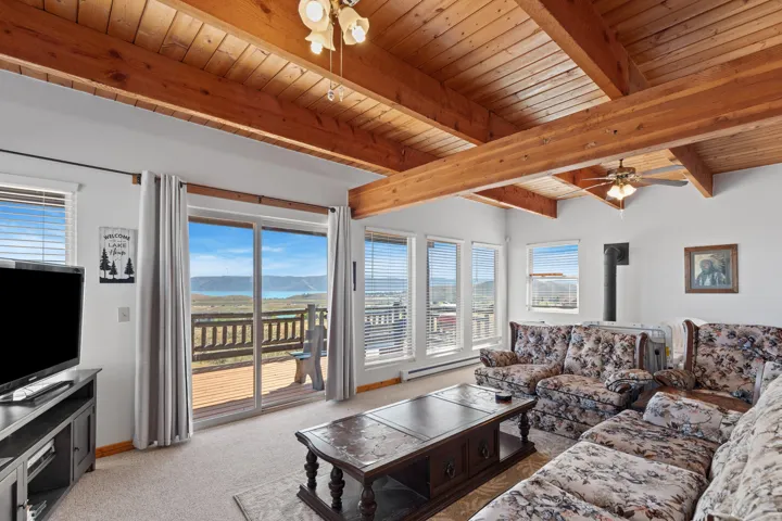 Cathedral wood-beam ceiling with exposed wood planks, large sliding glass door with exterior deck, multiple windows, built-in stove pipe, and recessed ceiling fan light fixtures
