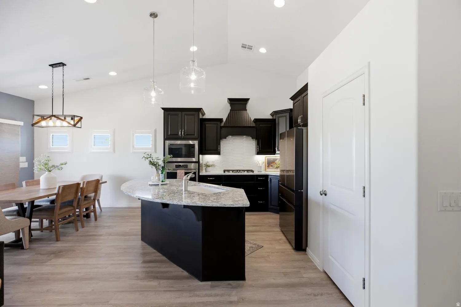 Vaulted ceilings with recessed lighting, wood-finish flooring, kitchen island with built-in sink and stone countertop, dark cabinetry, and integrated stainless steel appliances
