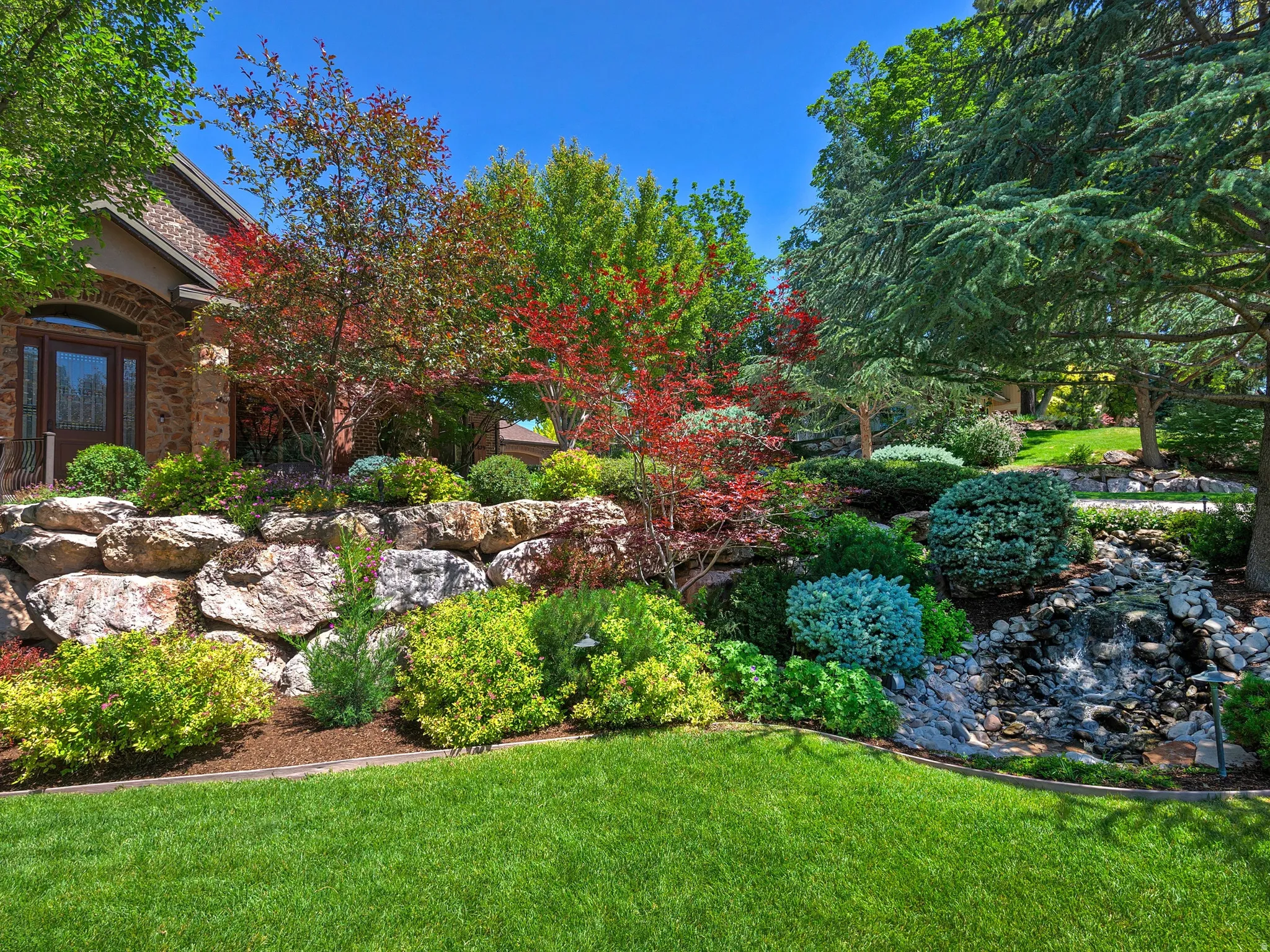 Stone retaining wall with varied plantings, manicured lawn, and lush landscaping, featuring a natural rock water feature