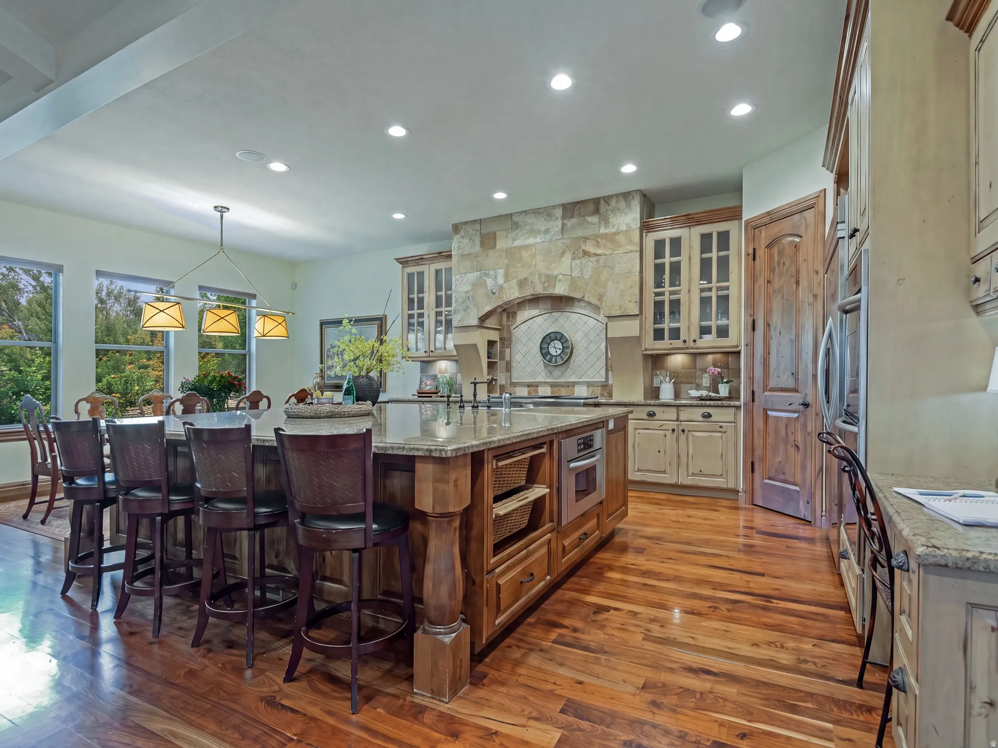 Open-concept kitchen with wood-finish flooring, a large island featuring integrated seating and storage, stainless steel appliances, and stone-clad range hood