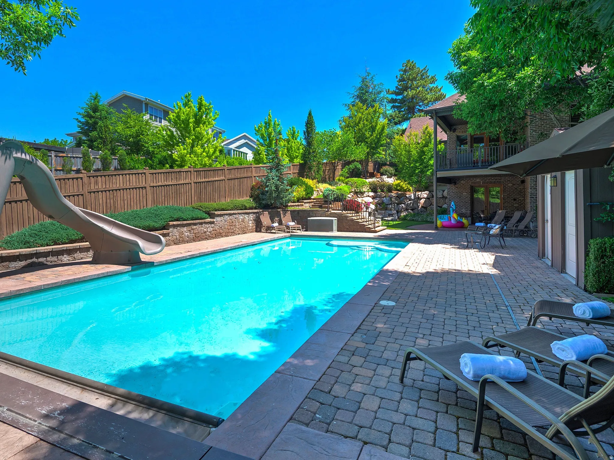 Outdoor swimming pool with integrated slide, surrounded by a paver patio and tiered landscaping, featuring a privacy fence