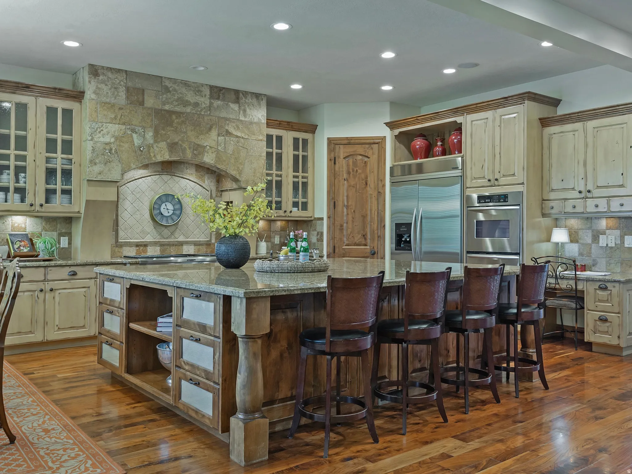 Expansive kitchen featuring a large island with seating, stainless steel appliances, stone-clad range hood, wood-finish flooring, and custom cabinetry