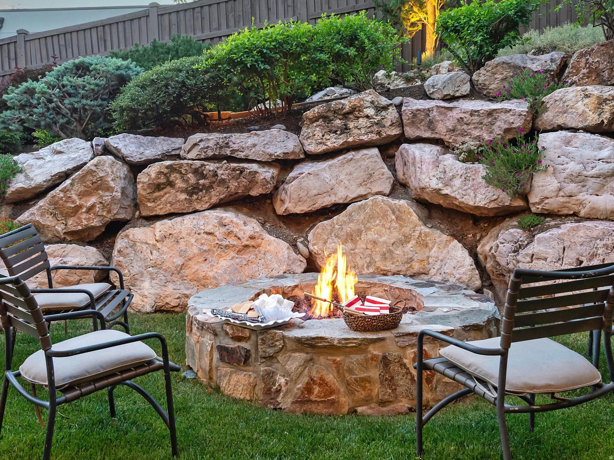 Stone fire pit with a natural rock retaining wall, surrounded by green lawn and established plantings