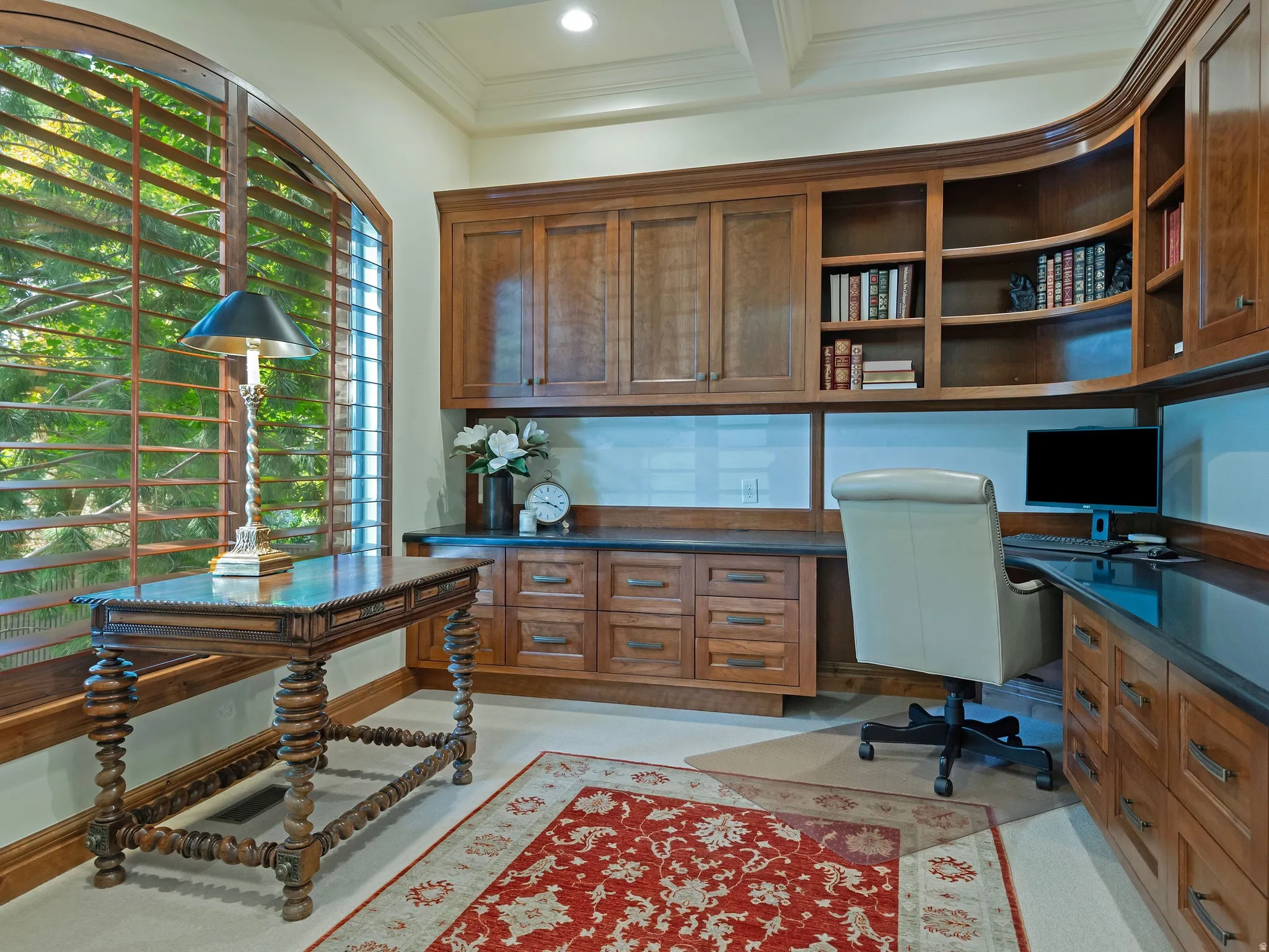 Arched window with wood plantation shutters, custom built-in wood cabinetry, integrated desk space, display shelving, and dark-toned countertops