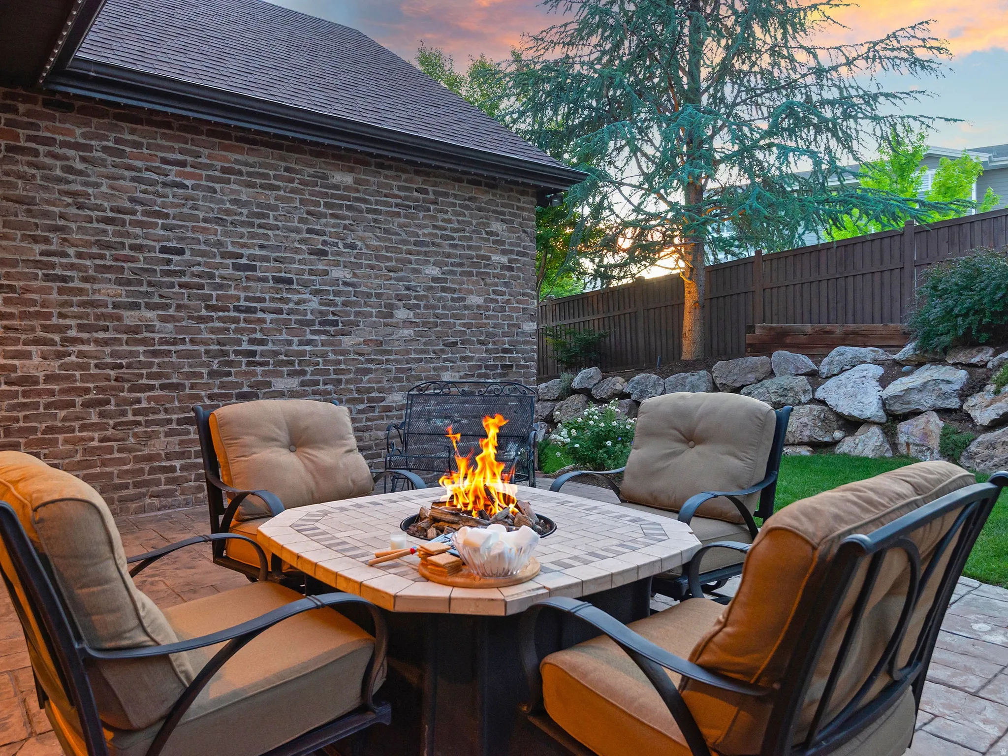 Paved patio with a built-in fire pit, brick exterior, and a rock retaining wall with established plantings