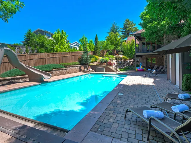 Outdoor swimming pool with integrated slide, surrounded by a paver patio and tiered landscaping, featuring a privacy fence