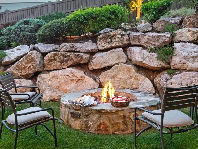 Stone fire pit with a natural rock retaining wall, surrounded by green lawn and established plantings