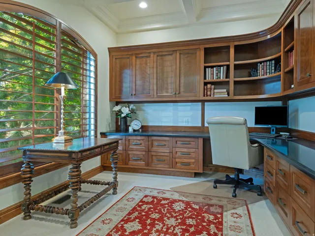 Arched window with wood plantation shutters, custom built-in wood cabinetry, integrated desk space, display shelving, and dark-toned countertops
