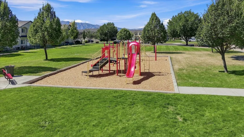 Expansive green space featuring a playground with slides and climbing structures, mature trees, a park bench, and a concrete walkway