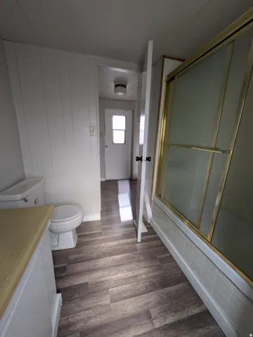 Wood-finish flooring with white baseboards, white paneled walls, and a shower enclosure with a gold-tone frame