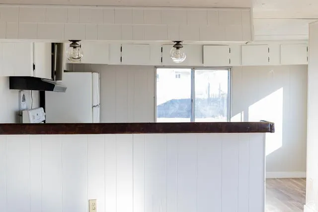 Modern kitchen featuring white shaker-style cabinetry, stainless steel appliances including a refrigerator, dark wood-finish countertops, pendant lighting with glass shades, and large windows framing natural light and distant landscape views