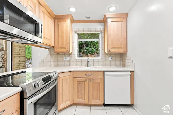 Kitchen featuring light wood cabinetry, light-toned countertops, a stainless steel microwave, a built-in dishwasher, and a tile backsplash