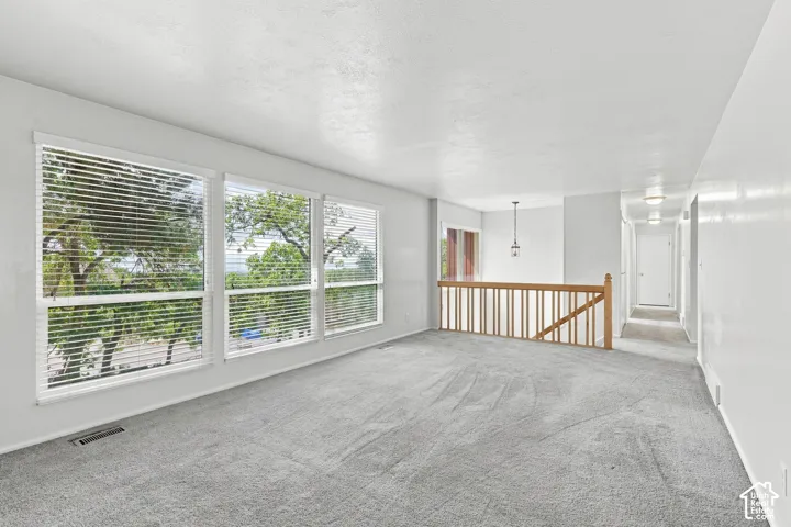 Large windows with integrated blinds, light gray carpeting, wooden railing, and a hallway with flush-mount lighting fixtures