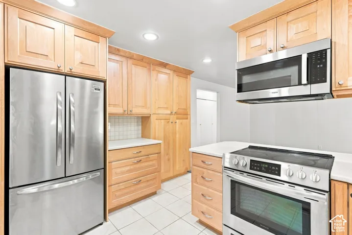 Kitchen featuring stainless steel appliances, light wood-finish cabinetry, white countertops, tile flooring, and a tile backsplash