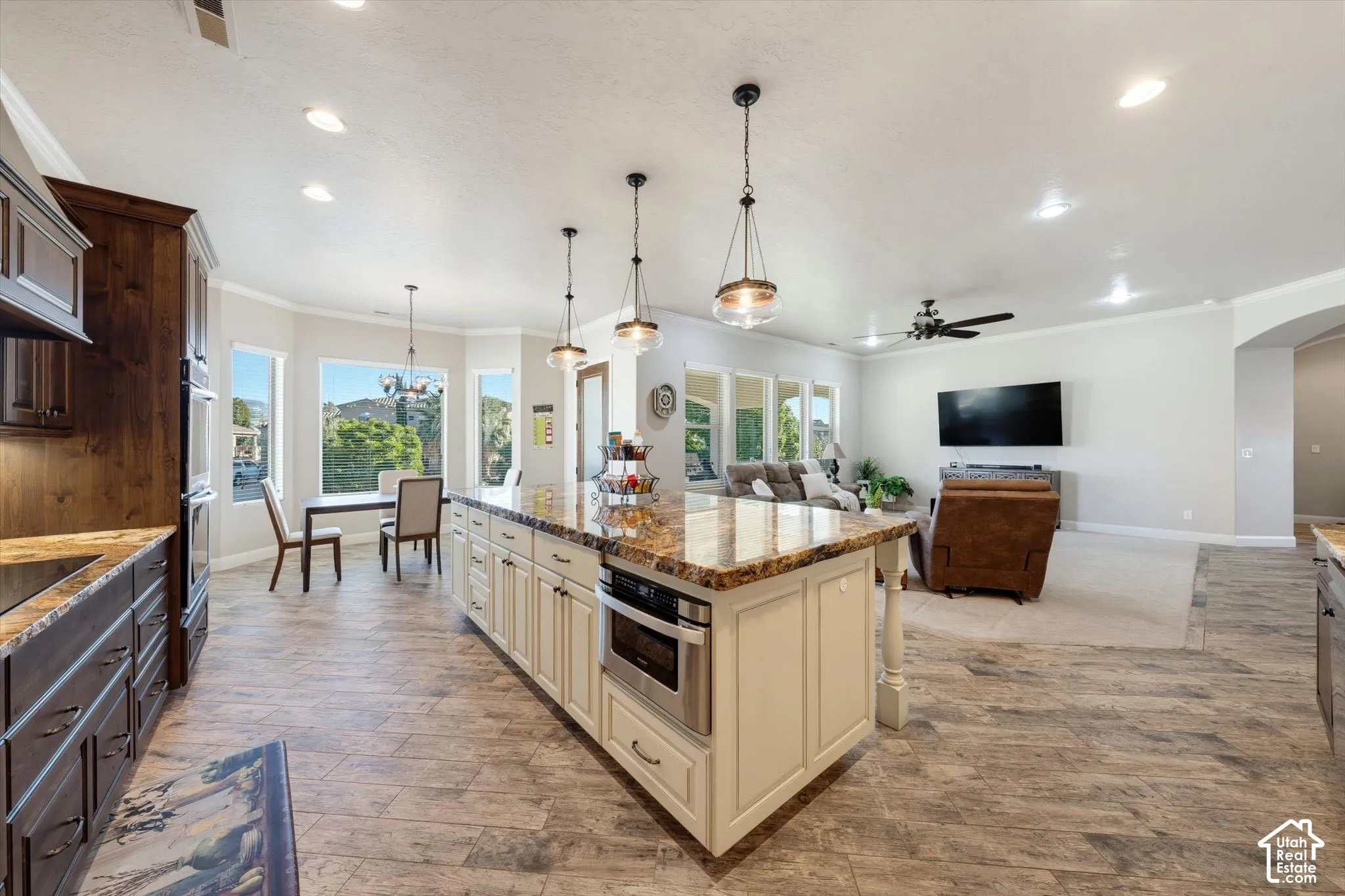 Open-concept kitchen and living area featuring a large center island with cream cabinetry and stone countertops, stainless steel built-in appliances, wood-finish flooring throughout, pendant lighting, and abundant natural light from multiple windows and g