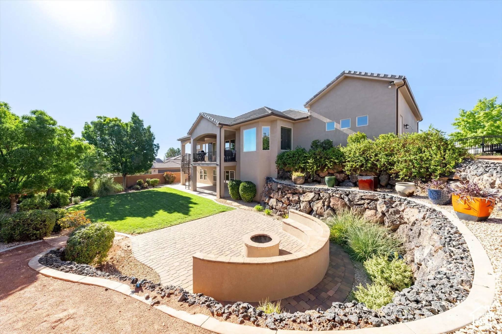 Expansive backyard featuring a manicured lawn, multi-level hardscaping, a paver patio with a built-in fire pit, and a covered balcony