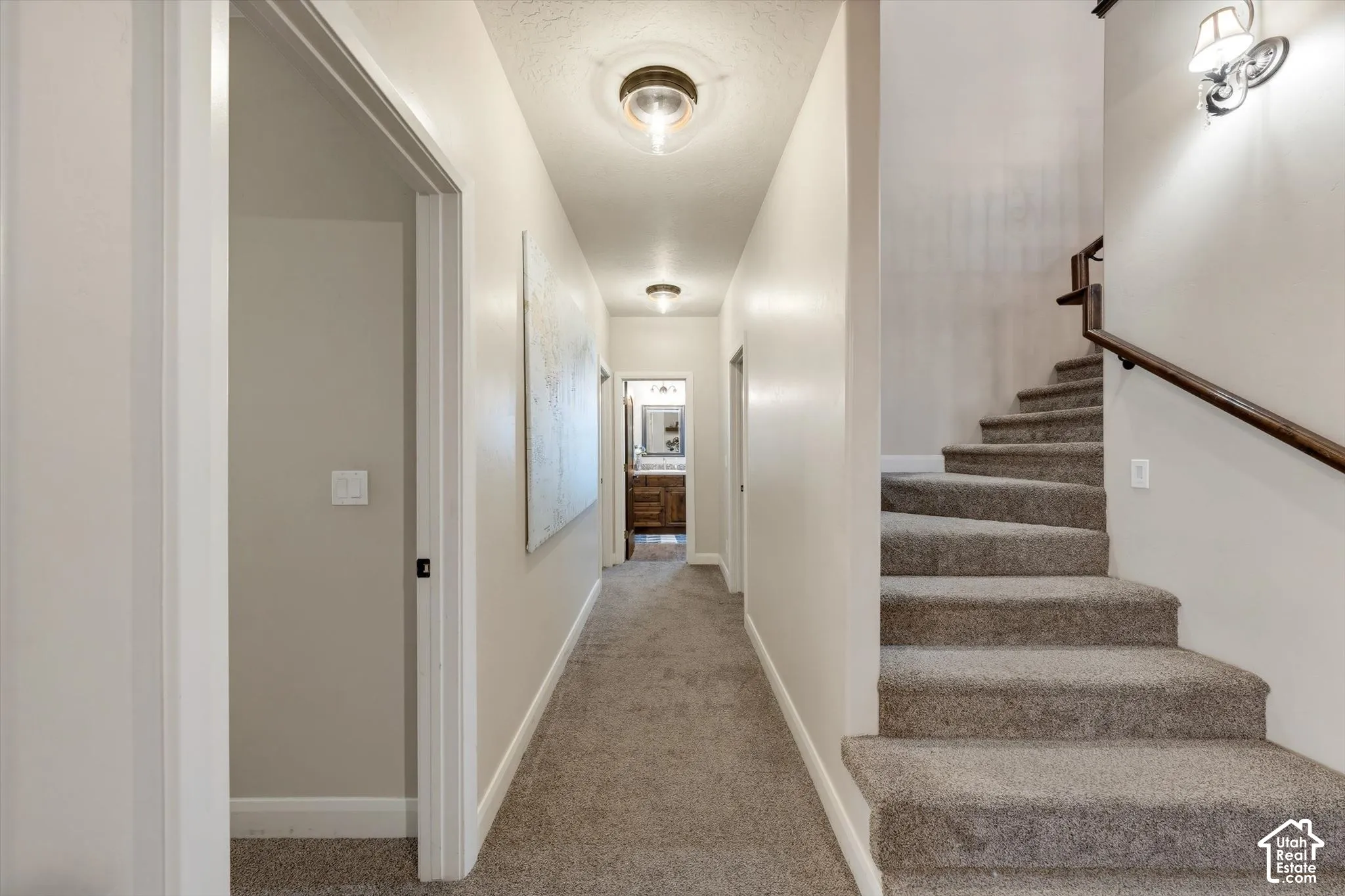 Carpeted staircase with dark wood-finish handrail, leading to a hallway with recessed lighting and a visible room with a wood-finish vanity