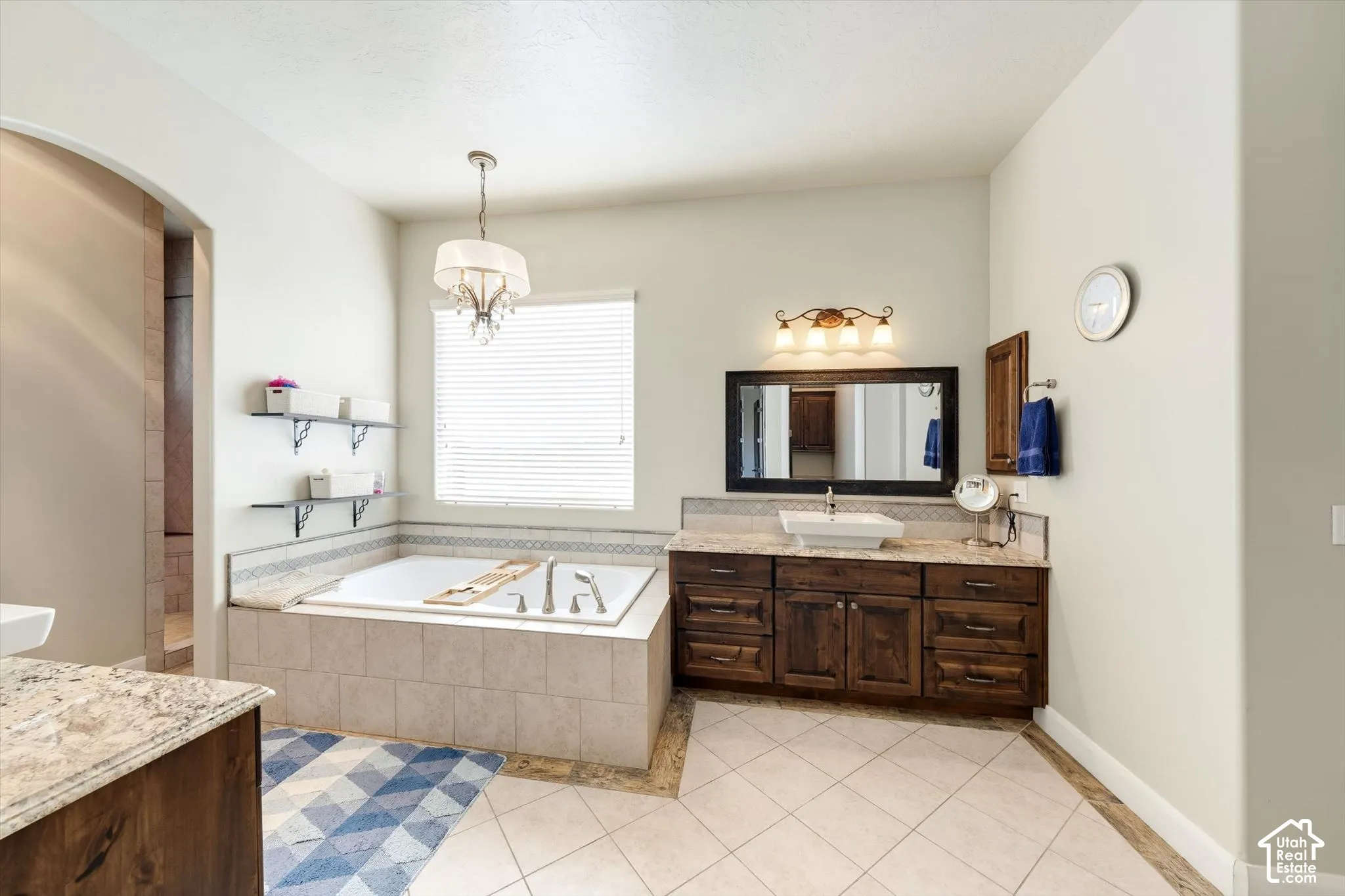 Soaking tub with tiled surround, wood-finish vanity with vessel sink, and an arched doorway