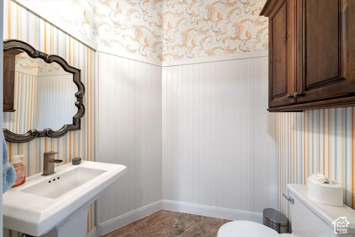 Pedestal sink with a bronze-finish faucet, an ornate mirror, wood-finish flooring, white beadboard wainscoting, and an upper wood cabinet