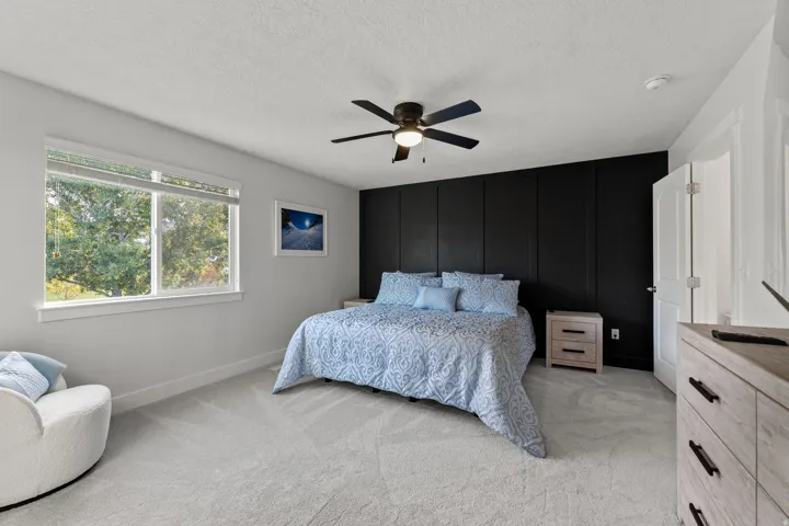 Bedroom featuring a paneled accent wall, light-colored carpeting, a ceiling fan with integrated lighting, and a window with blinds