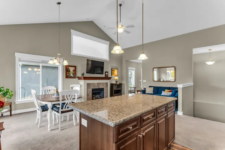 Vaulted ceiling with ceiling fan, island with granite countertop and rich wood-finish cabinetry, built-in fireplace with tile surround and wood mantel, large window with white trim, and recessed lighting