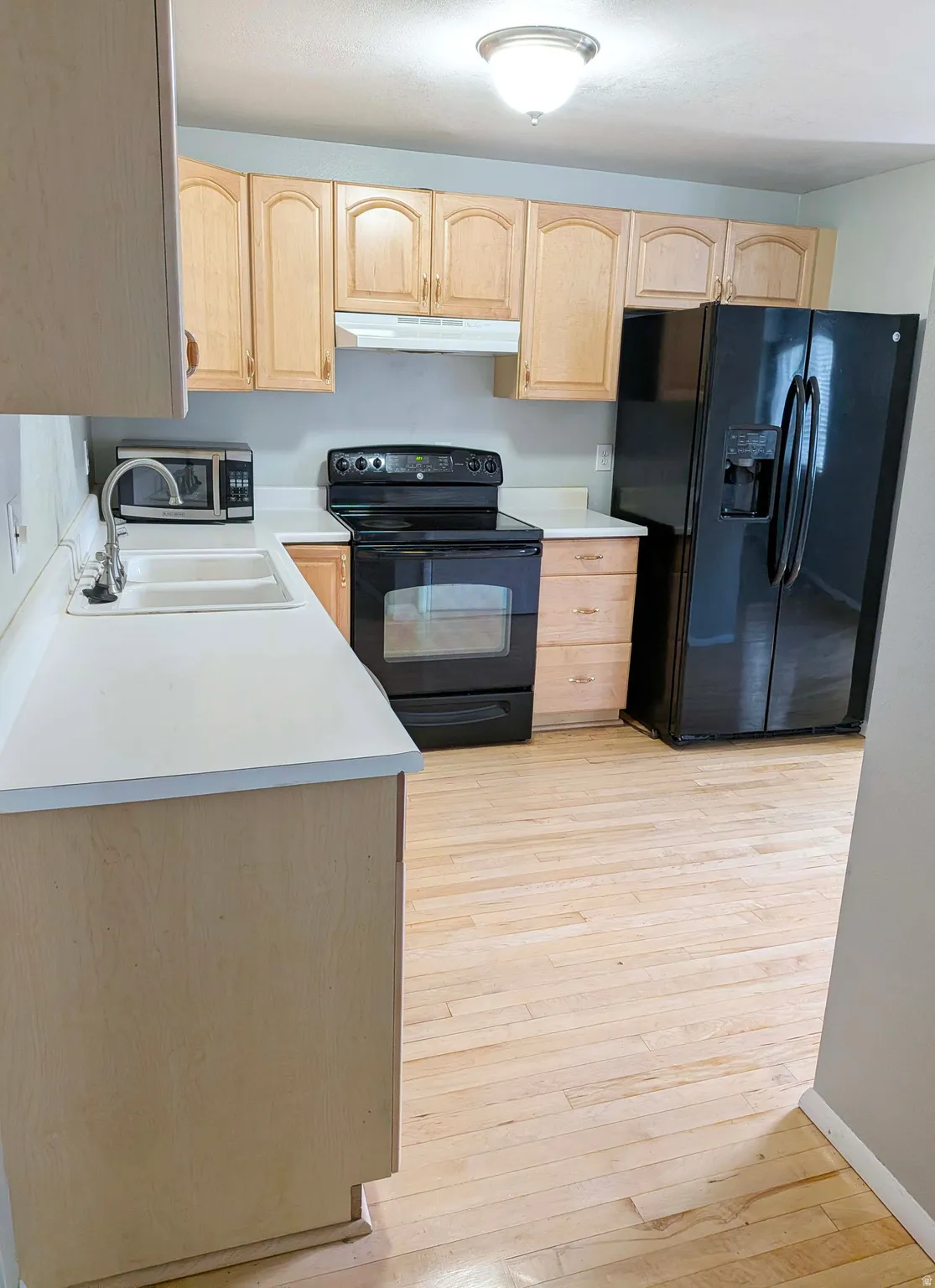 Spacious kitchen featuring wood cabinetry with arched door detailing, wood-finish flooring throughout, black side-by-side refrigerator, black electric range with oven, white undermount sink, and ample counter space with a large island