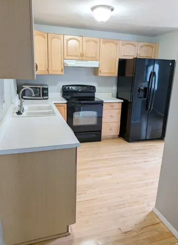 Spacious kitchen featuring wood cabinetry with arched door detailing, wood-finish flooring throughout, black side-by-side refrigerator, black electric range with oven, white undermount sink, and ample counter space with a large island