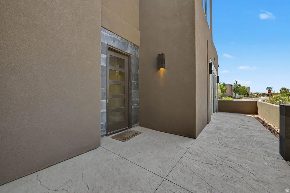 Modern architectural exterior featuring clean concrete pavement, stucco facade with warm neutral tones, recessed lighting, and a contemporary metal-frame entry door with frosted glass panels